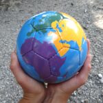Zwei Hände halten einen Fußball, der als Weltkugel bemalt wurde