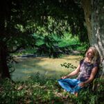 Frau sitzt meditierend an einen Baum gelehnt am Ufer eines Flusses