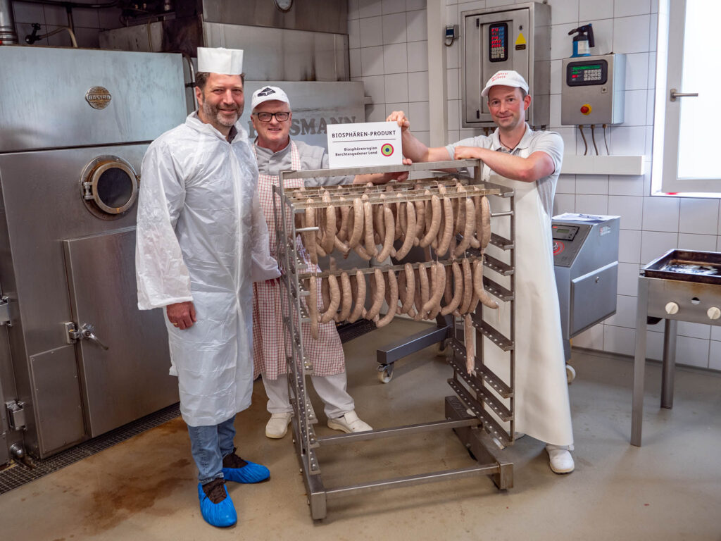 Die Metzgermeister Sichert und Braunsperger zusammen mit Dr. Peter Loreth, Leiter der Verwaltungstelle der Biosphärenregion, mit den für den Räucherprozess vorbereiteten Biosphären-Kabanossi und Biosphären-Produkt-Schild