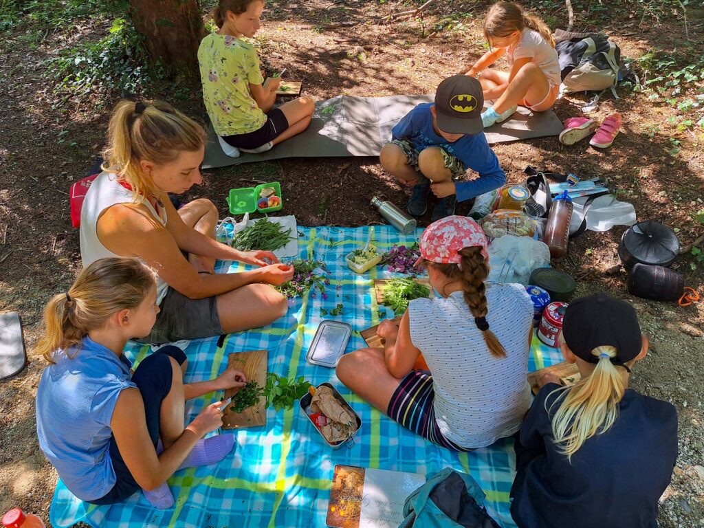 Kinder sitzen mit Mitarbeiterin der Biosphären-Verwaltungsstelle auf einer Picknickdecke im Wald und befassen sich mit gepflückten Wildkräutern