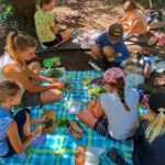 Kinder sitzen mit Mitarbeiterin der Biosphären-Verwaltungsstelle auf einer Picknickdecke im Wald und befassen sich mit gepflückten Wildkräutern