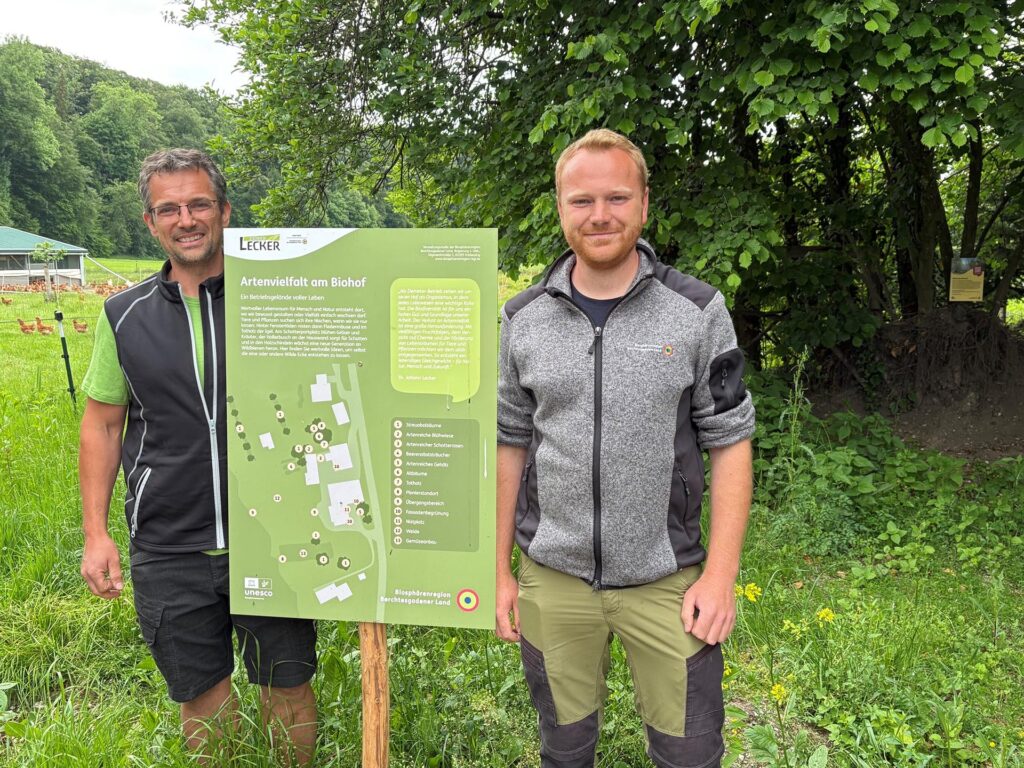Hans Lecker vom Biohof Lecker und Luis Frank von der Biosphären-Verwaltungsstelle mit einem Schild zum Thema "Wilde Ecken"